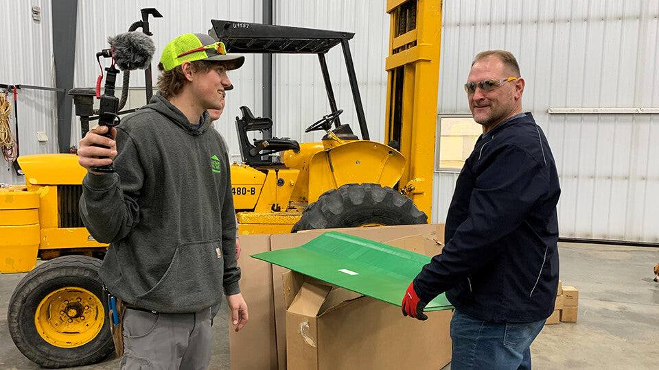 Load video: Chet Larson of Larson Farms holding a camera and smiling next to Eric from May Wes Manufacturing holding green poly liners for grain head auger trough liner installation.