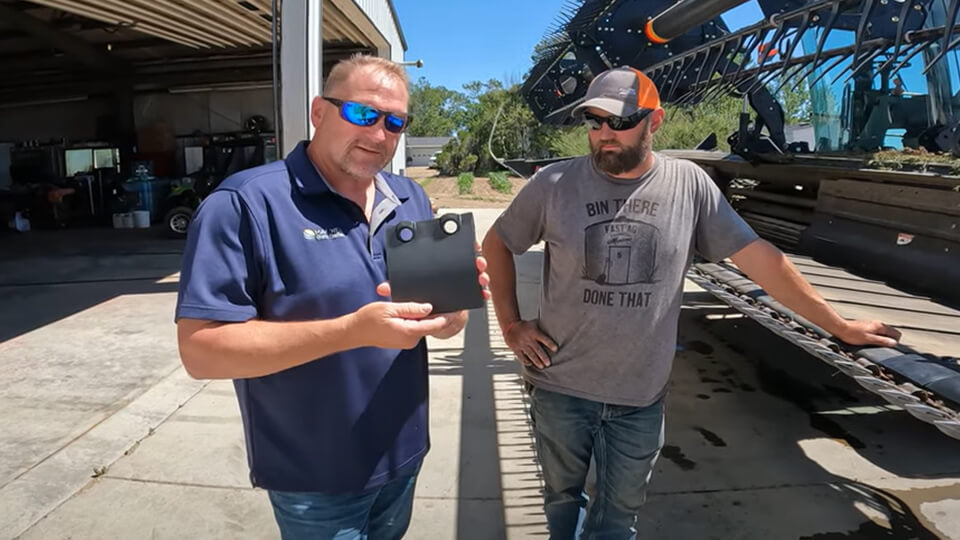 Eric from May Wes Manufacturing holding molded cutterbar skid shoe for MacDon, standing next to Tony Fast of Fast Ag Montana in front of MacDon grain head.